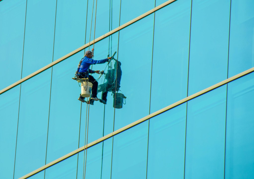 Window cleaner suspended on ropes wiping a bright blue high-rise glass facade, buckets hanging beside him