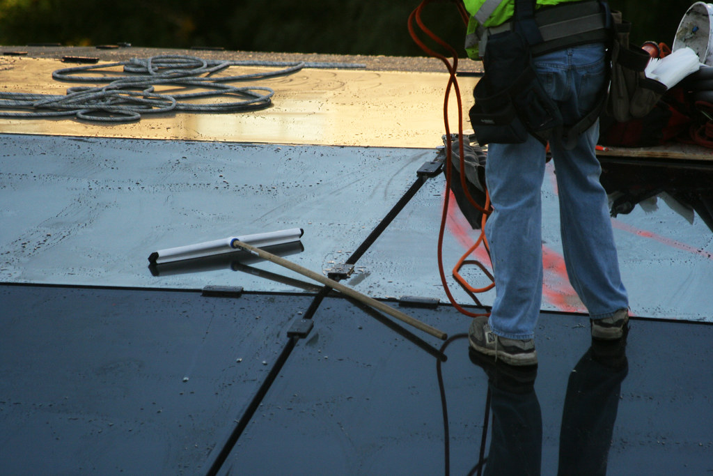 Worker in jeans and a hi-vis vest pulling a squeegee across wet glass roof panels, ropes coiled nearby