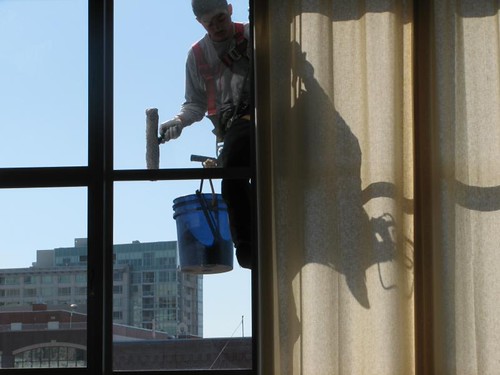 Window cleaner with a blue bucket scrubbing a tall multi-pane window beside a curtain, city buildings behind