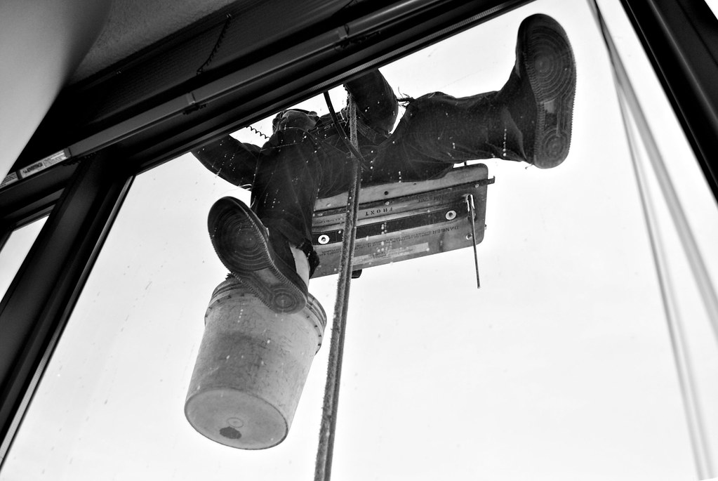 Black-and-white close view of a rope-access cleaner's boots, bucket and bosun's chair against bright glass