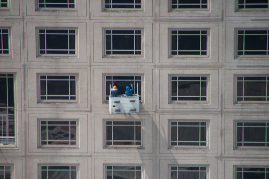 Two window cleaners on a suspended platform halfway up a concrete office facade, row upon row of windows around them