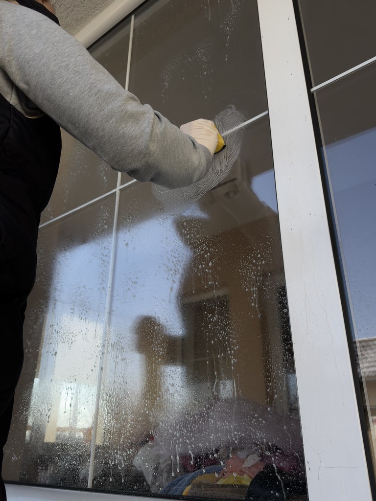 Cleaner in a grey sweatshirt scrubbing a tall interior glass door with a yellow sponge in daylight