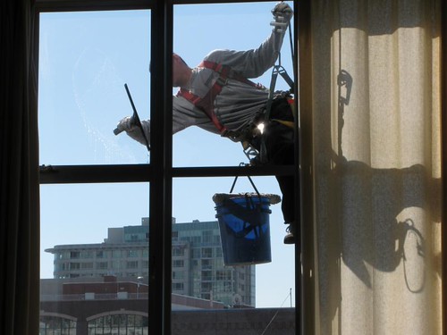 Rope-access window washer silhouetted against blue sky, bucket hanging from harness, city buildings behind