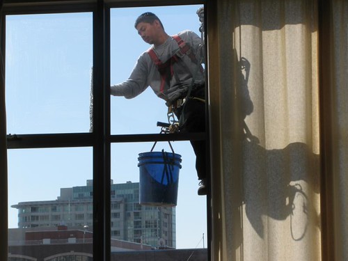 Rope-access window cleaner in harness and red straps wiping an exterior window, blue bucket at his side