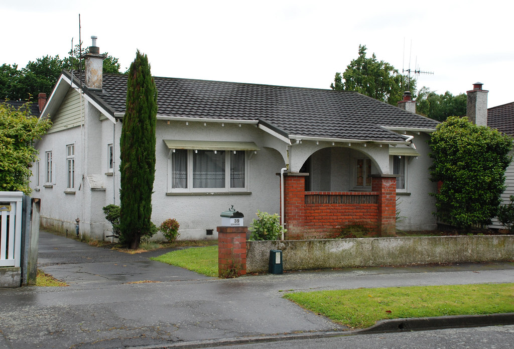 White stucco New Zealand suburban bungalow with brick porch, tile roof and front lawn on an overcast day