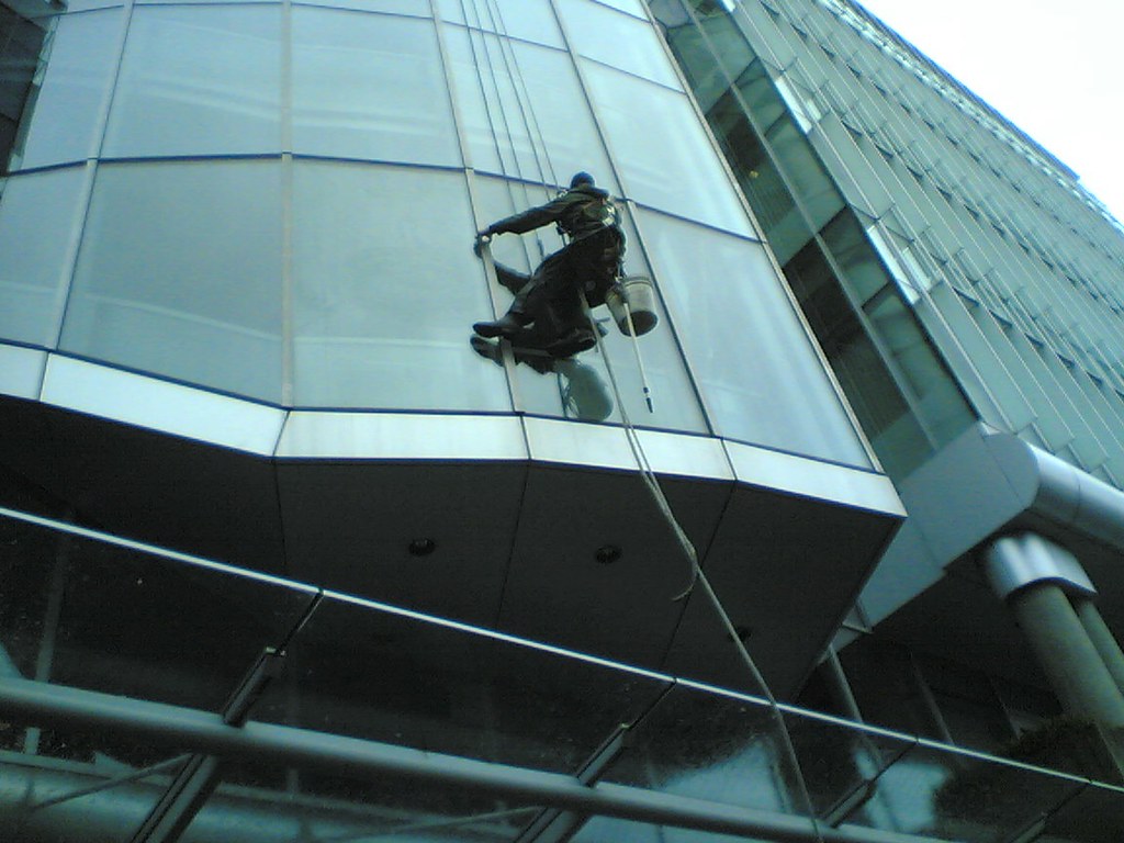 Abseiling window cleaner with a bucket working down a modern blue-glass office facade, viewed from below