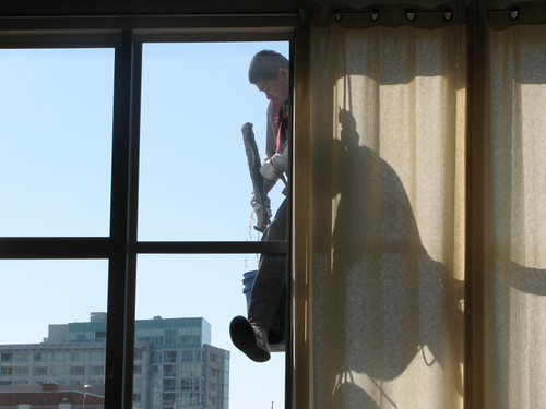 Silhouette of a rope-access washer with tools at a tall window, seen from inside past a sheer curtain
